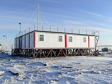 Zu sehen ist ein Containerbau auf Holzstelzen in einer schneebedeckten Landschaft.