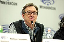 A man with greying brown hair, glasses, a vest, and a striped button-up shirt speaks at into a microphone.