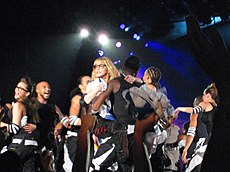 Medium range shot of a group of people, wearing black-and-white colored dress on stage. Central to them is a blond woman, wearing black bordered glasses, being held up by a man. Lights fall on the stage from above.