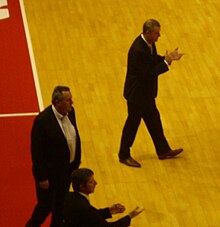 entraîneur rentrant sur le terrain en applaudissant. Deux autres hommes en costume sont au bord du terrain.