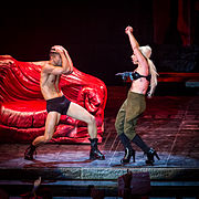 Lady Gaga singing while wearing a rifle-equipped bra, accompanied by a male dancer who is wearing a black underwear and boots. In the background a couch is visible, which looks like it was made from meat.