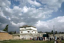 Un bâtiment carré de deux étages derrière un mur ; au premier plan un attroupement en costume pakistanais regarde la maison.