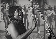Black-and-white photograph of a black woman singing into a microphone, with a grainy photograph of various people on a street visible in the background.