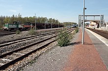 Train de wagons-trémie (avec deux locotracteurs à sa tête), voies, quais et passerelle, vus par temps ensoleillé.