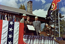 A large house with a stone chimney, decorated with bunting. An army officer runs a red and blue flag up a flagpole. In the foreground are two men in suits and one in an army uniform.