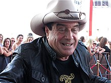 Upper body shot of a smiling man in a cowboy hat and black leather jacket. He is wearing a black T-shirt with a gold design which is mostly out of shot. In the background are people behind a barrier fence.