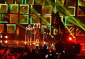 Three women wearing red and black dresses are seen performing to a crowd in front of an LED background displaying green boxes.