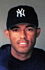 A headshot of Mariano Rivera smiling while wearing a midnight blue baseball cap and baseball jersey in front of a concrete backdrop.