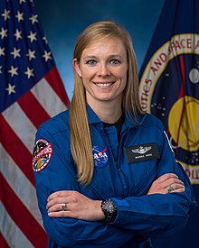 A woman in a blue flight suit is sitting in front of US & NASA flags; she is facing the camera and smiling.