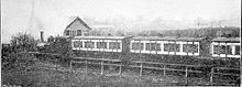 A black and white photograph showing a small train pulling into a small, rural railway station.