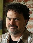 A portrait of a man with curly black hair and a salt-and-pepper beard facing the left of the screen, eyes turned towards the viewer