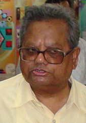 Colour closeup photograph of a man wearing rectangular-framed spectacles with receding grey hairline