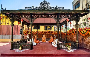 Sculptures of five people clad in traditional Indian clothing sitting under an ornate black canopy in a semicircle on the floor around a sculpture of a bespectacled person in saffron clothes.