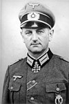 The head and shoulders of a man. He wears a peaked cap and a military uniform and an Iron Cross displayed at the front of his uniform collar. His facial expression is determined; his eyes are looking into the camera.