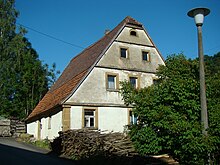 Nordwest- und Südwestwand der Dorfmühle Horrenbach. Ein hier auf der Hangseite einstöckiges Gebäude mit einem drei Stockwerke hohen Giebel mit Krüppelwalmdach. Das Gebäude ist weiß gestrichen mit Tür- und Fensterrahmen aus ockerfarbenem Sandstein. Im Umfeld des Gebäudes sind mehrere Brennholzstapel, am Bildrand Bäume, Büsche und eine Straßenlaterne.