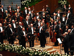 Several dozen musicians in formal dress, holding their instruments, behind a conductor