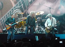 Led Zeppelin onstage: John Paul Jones playing bass guitar, Robert Plant holding a microphone, and Jimmy Page playing guitar.