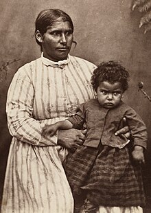 Seated Aboriginal woman in a striped dress holding a child on her lap