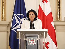 A woman with dark hair and a white suit jacket speaks behind a podium and in front of a blue NATO flag and red and white Georigan flag.
