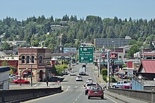Looking down from a one-way bridge with two lanes at a city skyline with low-slung buildings and hills in the background; a sign reading "101 North Hoquiam, Port Angeles; 12 East Olympia" hangs over the highway.