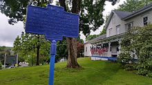 a picture of thee marker, instead of yellow letters on blue the marker is all blue, in the background you can see the wood house with flags and that there is a sign saying it is a Masonic lodge