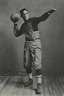 Pollard in football uniform staring off in the distance and striking a classic football pose; one arm is outstretched, while the other holds a football, as if he were about to throw it.