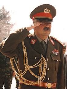 Photograph of Abdul Rashid Dostum wearing a black suit with a lapel pin, white shirt, and necktie