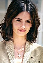 A close-up of a female with dark brown hair roots, and the rest of her hair is medium brown. Her hair is short and the bottom of her hair is curly. She is wearing a small cross necklace on her neck and a tan colored shirt that has a long shirt collar.