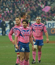 Photographie en couleurs. Quatre joueurs portant un maillot rose et bleu sont debout, avec Christophe Dominici en premier plan, regardant vers la gauche les sourcils froncés.