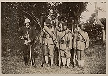 un officier en uniforme bleu foncé au côté de six tirailleurs malgaches en uniforme kaki