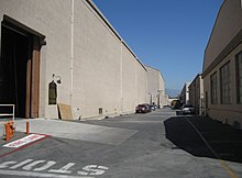 Bordering an asphalt road, receding into the distance, are two rows of large beige coloured buildings. Another beige building blocks the horizon, above which mountains are visible in the distance. The sky is blue with no clouds. Several cars are parked outside the buildings. Near the photographer, at a large open entrance to one building, the word STOP has been painted in white on the road. Next to it, a fire hydrant is visible, in front of which the words FIRE LANE are painted on the road.