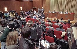 Salle pleine de personnes, assises sur des chaises. Au premier plan et sur la gauche, des cameramen et des photographes. En arrière-plan, 4 personnes leur faisant face, assises côte à côte sur une estrade.