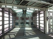View of the end of the Tribeca pedestrian bridge. There are four steel-and-glass doors in the center of the picture, marking the entrance to the school. The bridge is entirely enclosed by glass panes with metal beams on three sides, as well as a steel floor.