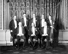 A monochrome photograph of men wearing white tie with breeches, three of them (Baldwin for the United Kingdom, George V, and King for Canada, from viewer's left to right) seated in the front row and five (Monroe for Newfoundland, Coates for New Zealand, Bruce for Australia, Hertzog for South Africa, and Cosgrave for Ireland, ditto) standing behind them.
