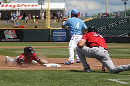 A man in a red baseball jersey and gray pants kneels down on a green grassy field as a player in the same uniform slides headfirst into third base.