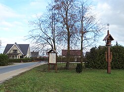 Image of a green lawn next to a street with a house in the background. On the lawn are three trees and a sign in front of the trees.