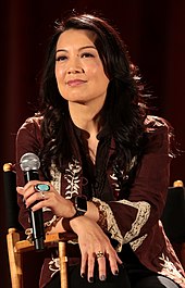 A woman in a patterned dress sits in a chair and holds a microphone while looking off-camera.