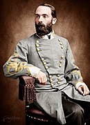 Colorized photo of a bearded man seated in a chair. He wears a gray military uniform with two rows of gold buttons and yellow frogging on the sleeves.