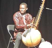 Sidiki Diabaté sitting on a chair onstage, playing a kora and looking right of camera