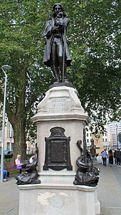 Larger than lifesize bronze statue of man in period clothes, standing with one hand on a staff, the other raised to his chin. It is on a white stone pedestal with inscription "Edward Colston Born 1636 Died 1721", and bronze inscribed plaques below. Large bronze dolphins are on each corner of the base. It is in an urban setting with a large tree behind and above it.