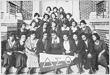 Un groupe de jeunes femmes afro-américaines, en uniforme, assises sur les escaliers d'un bâtiment en brique. Le rang de devant tient une banderole à l'effigie de la sororité.