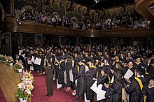 Young adults wearing ceremonial black robes and graduation caps stand at their seats in an ornate hall while onlookers in a three-sided balcony applaud above them.