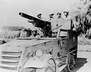 A photograph of an M3 gun motor carriage in a flat area. A mountain is visible in the background, but is obscured by clouds.