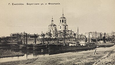 Photographie en noir et blanc de navires de la flotte fluviale dans le port de Ienisseïsk, avec au fond la cathédrale de l'Épiphanie et d'autres bâtiments.