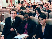 Photographie en couleur avec au premier plan deux femmes et un homme assis en rangée ; en arrière plan une multitude de personnes assises en rangées successives.