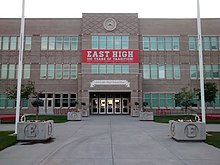 A long shot of the exterior of a three-storey public high school building with a banner reading "Salt Lake High School East". The exterior prominently features windows, as well as a large clock at the top of the entrance.