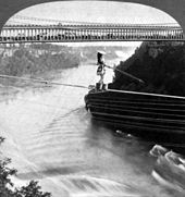 A woman is crossing on a rope over a river. She wears a wide-brim hat and holds a pole to balance herself while her feet are in buckets. A double-deck bridge, filled with an audience on the lower deck, is in the background.