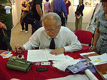 A man sits at a table with a book and a pen in hand