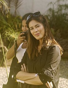 An image of Aquino wearing a black long-sleeved shirt with her arms crossed