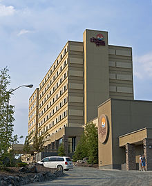A beige and tan concrete high-rise seen from the base of an upwardly sloping gravel driveway curving around a planted area. On the building, and the side of a pavilion projecting to front right, are circular decorative signs identifying it as the Explorer Hotel.